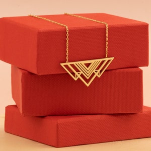 May include: A gold necklace with a geometric triangle pendant design. The necklace is displayed on a stack of three red boxes.