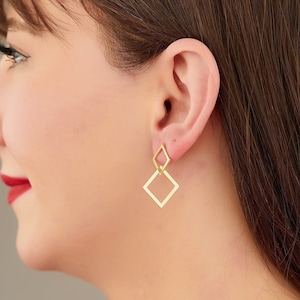 May include: A woman with long brown hair wearing gold geometric earrings. The earrings are made of two connected squares.