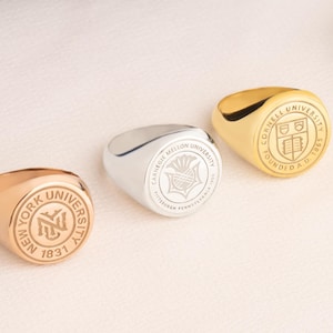 May include: Three signet rings in gold, silver, and rose gold, each with a university crest. The rings are displayed on a white surface with a wooden rainbow arch and greenery in the background.