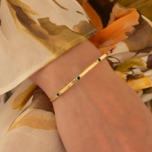 May include: A gold bracelet featuring three small, green gemstones. The bracelet is worn on a wrist, against a backdrop of a floral blouse in yellow, orange, and green. The bracelet's design is simple and elegant, suitable for everyday wear.
