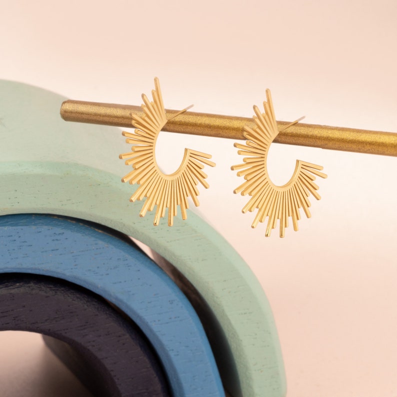 Peut inclure: Une paire de boucles d'oreilles cr&eacute;oles dor&eacute;es avec un motif soleil. Les boucles d'oreilles sont pr&eacute;sent&eacute;es sur un support en bois.