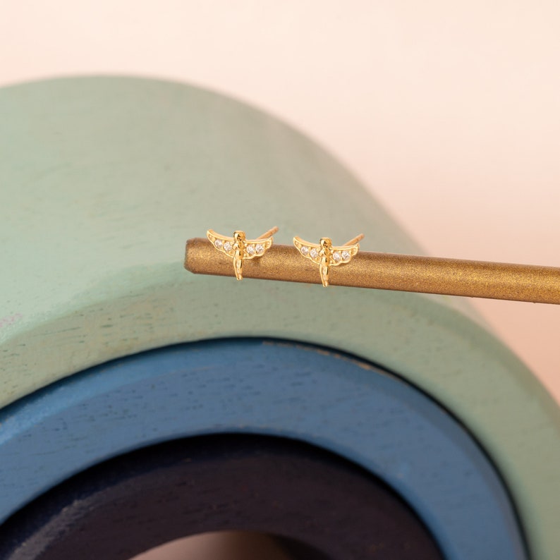 May include: A pair of gold dragonfly earrings with clear cubic zirconia stones. The earrings are on a gold surface.