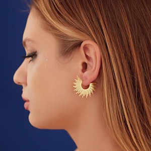 Peut inclure: Une femme aux longs cheveux blonds portant des boucles d'oreilles cr&eacute;oles dor&eacute;es avec un motif soleil. Les boucles d'oreilles sont faites de fils m&eacute;talliques fins qui rayonnent du centre du cercle.