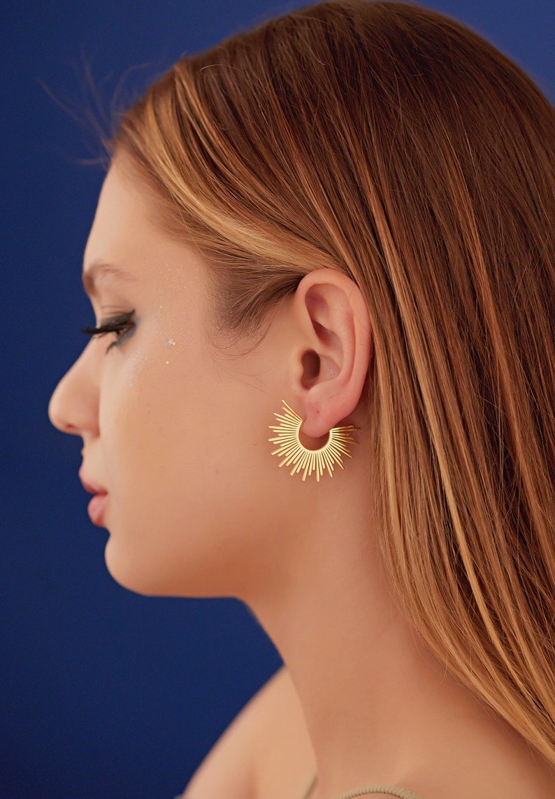 Peut inclure: Une femme aux longs cheveux blonds portant des boucles d'oreilles cr&eacute;oles dor&eacute;es avec un motif soleil. Les boucles d'oreilles sont faites de fils m&eacute;talliques fins qui rayonnent du centre du cercle.