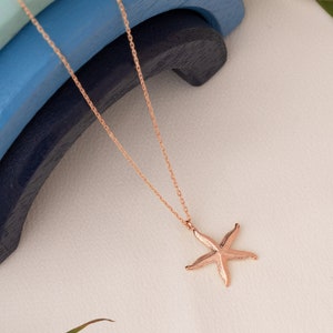 May include: A rose gold necklace with a delicate chain and a starfish pendant. The necklace is displayed on a white surface with blue and green wooden blocks in the background.