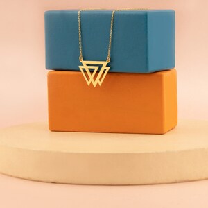 Puede incluir: Un collar dorado con un colgante geométrico de triángulo. El colgante presenta tres triángulos interconectados. El collar se muestra sobre un bloque azul y naranja, sobre un fondo rosa claro.