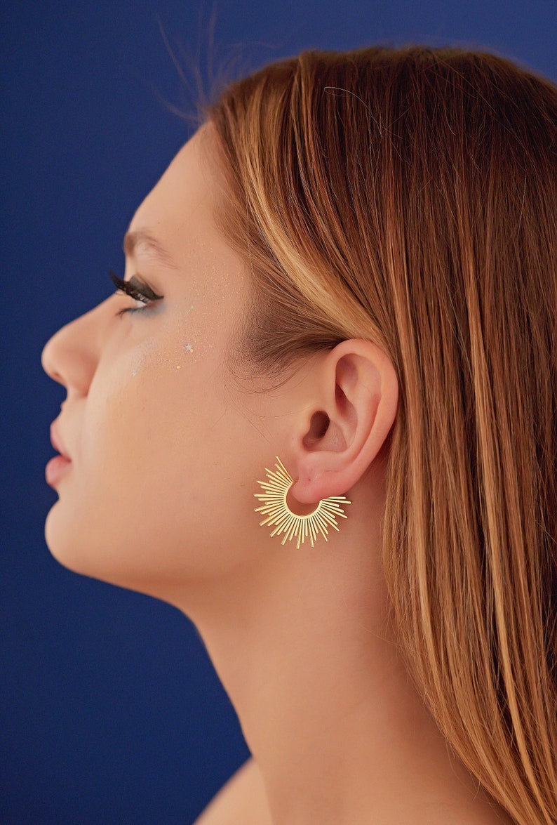 Peut inclure: Une femme aux longs cheveux blonds portant une paire de boucles d'oreilles cr&eacute;oles dor&eacute;es avec un motif soleil.