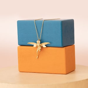 May include: A gold necklace with a winged fairy pendant. The necklace is displayed on top of two blocks, one blue and one orange.