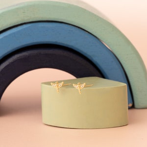 May include: A pair of gold earrings with a small angel design and clear crystal accents. The earrings are displayed on a green surface.