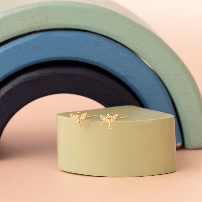 May include: A pair of gold earrings with a small angel design and clear crystal accents. The earrings are displayed on a green surface.