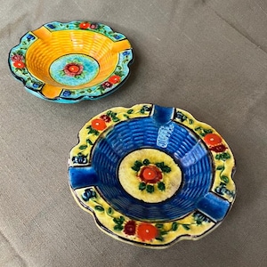 May include: Two decorative ceramic ashtrays. One is yellow and turquoise with red floral accents. The other is blue with yellow trim and red floral accents. Both have a circular shape.
