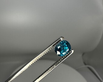 May include: A teal blue gemstone held in a pair of tweezers. The gemstone is oval shaped and has a faceted cut.