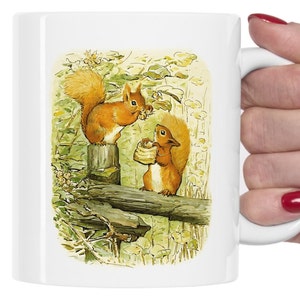May include: A white ceramic mug with a colorful illustration of two red squirrels on a wooden fence. The squirrels are holding nuts in their paws.
