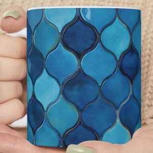 May include: A white ceramic mug with a blue and turquoise geometric pattern. The pattern resembles a mosaic of overlapping teardrop shapes.