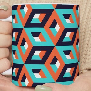 May include: A white ceramic mug with a geometric pattern. The pattern features orange, teal, white, and navy blue squares.