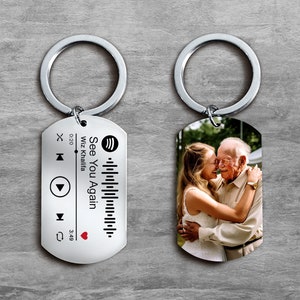May include: Two silver keychains with a photo of an older man and a younger woman hugging on one keychain and a music player design with the text "See You Again" and "Wiz Khalifa" on the other keychain.