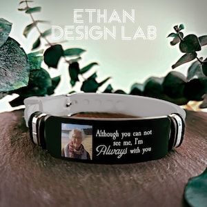 Può includere: Un braccialetto nero con una foto in bianco e nero di una donna anziana incisa e il testo "Although you can not see me, I'm Always with you". Il braccialetto è bianco e ha accenti argentati.