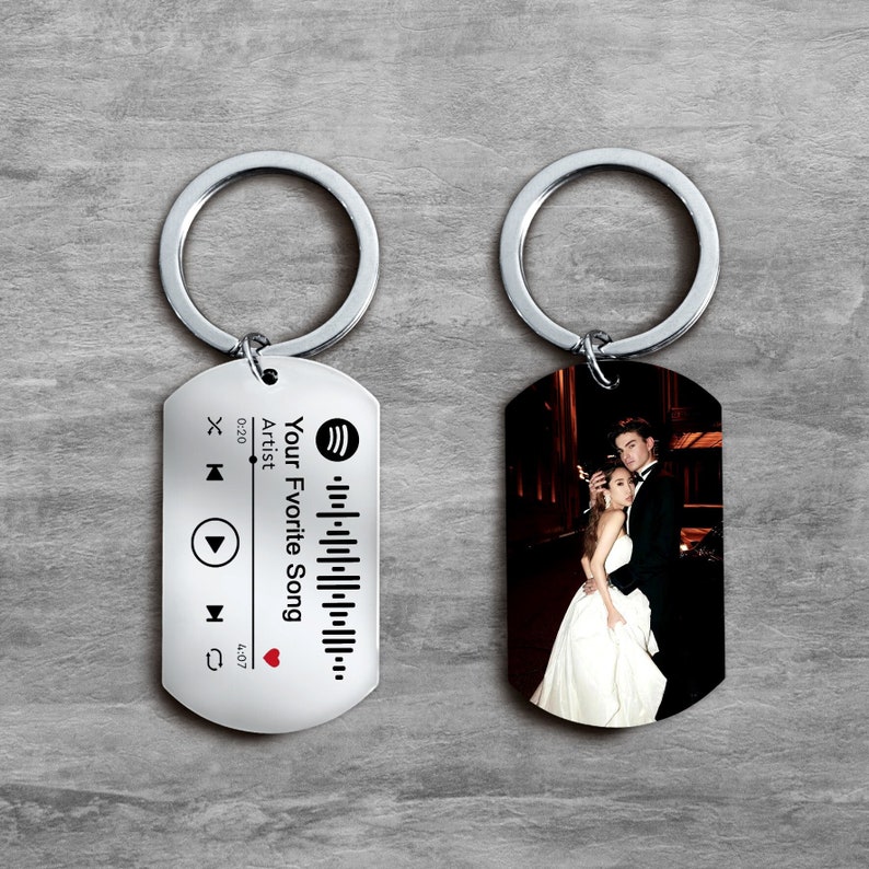 Puede incluir: Dos llaveros plateados con una foto en blanco y negro de una pareja en un llavero y una etiqueta plateada con un logotipo de Spotify, un c&oacute;digo de barras y el texto "Your Favorite Song" en el otro llavero.