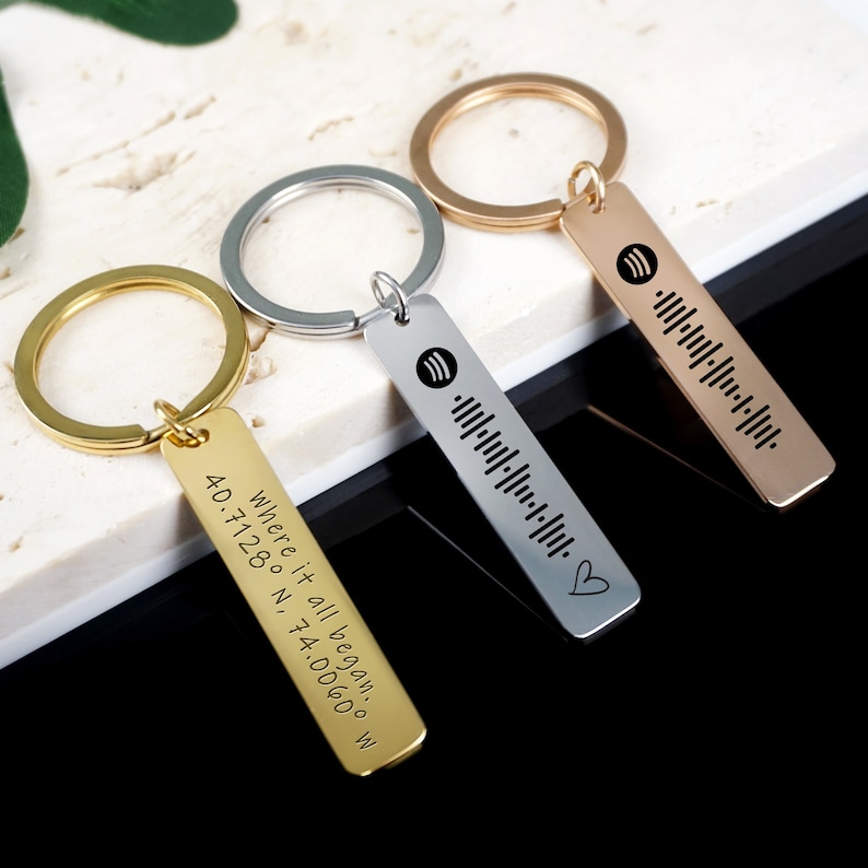 May include: Three gold, silver, and rose gold keychains with engraved bar pendants. The gold pendant has the text "Where it all began: 40.7128&deg; N, 74.0060&deg; W". The silver and rose gold pendants have a Spotify logo and a barcode.