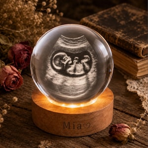 May include: A clear glass sphere featuring a laser-etched 3D ultrasound image of a foetus inside. The sphere rests on a circular wooden base that emits a warm light, with the name Mia and a heart symbol engraved on the front of the wooden stand.