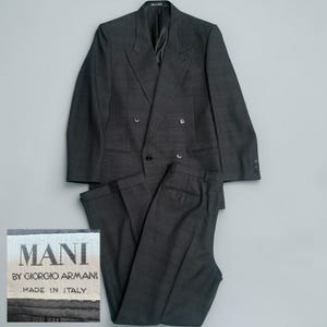May include: A charcoal gray double-breasted suit with matching trousers. The suit features a classic notched lapel and multiple buttons. The label reads "MANI BY GIORGIO ARMANI MADE IN ITALY."