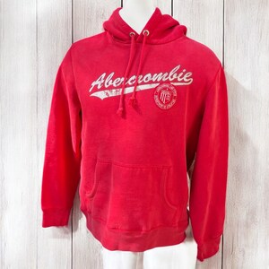 May include: A red Abercrombie hoodie with a drawstring hood and a kangaroo pocket. The white text "Abercrombie" is across the chest, with a circular logo on the right side. The hoodie is displayed on a mannequin.