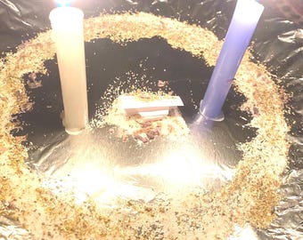May include: Two lit candles, one white and one purple, with bright flames, are positioned on a dark surface. The candles are surrounded by a circle of herbs and other materials, suggesting a ritual or spiritual practice.