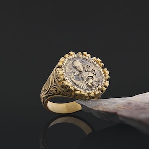 May include: A gold ring with a detailed design featuring a woman's face on a round coin-shaped top. The ring has a textured band with intricate floral patterns.