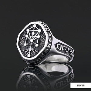 Op de afbeelding: Zilveren ring met een gedetailleerd ontwerp. De ring heeft een vierkante voorkant met een geometrisch patroon en een gegraveerde rand. De band heeft gegraveerde tekst. De ring wordt getoond op een reflecterend oppervlak.