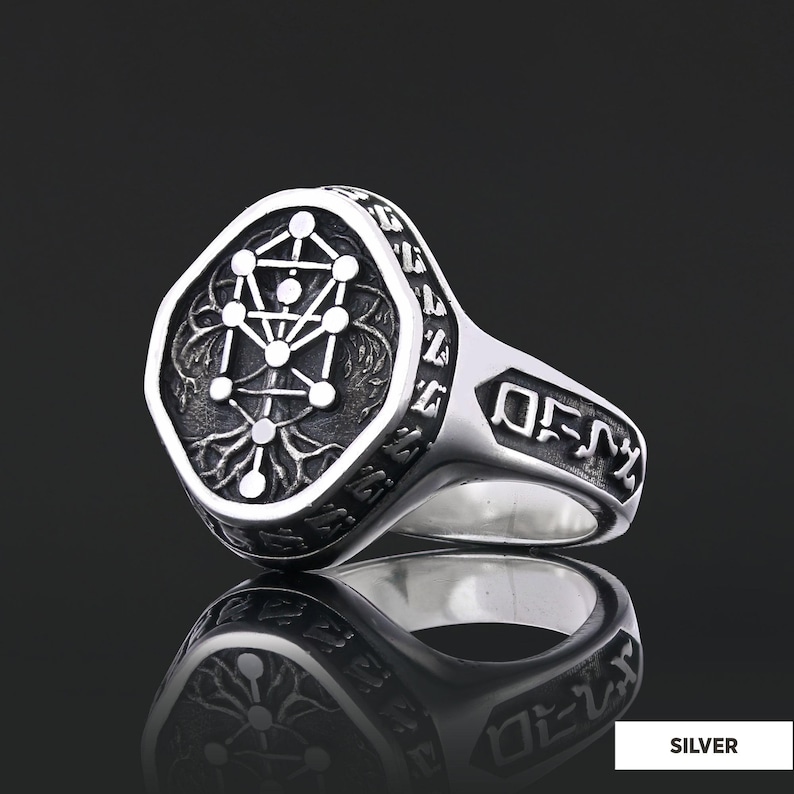 Op de afbeelding: Zilveren ring met een gedetailleerd ontwerp. De ring heeft een vierkante voorkant met een geometrisch patroon en een gegraveerde rand. De band heeft gegraveerde tekst. De ring wordt getoond op een reflecterend oppervlak.