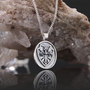 May include: Silver pendant necklace featuring a shield with a cross design. The shield is oval shaped and has a textured surface. The cross is detailed with floral accents.