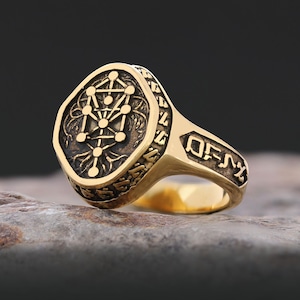 Op de afbeelding: Een goudkleurige ring met een ingewikkeld ontwerp. De ring heeft een gedetailleerd, verhoogd patroon op de voorkant, met een geometrisch symbool in het midden. De band heeft ge&euml;tste symbolen. De ring ligt op een stenen oppervlak.
