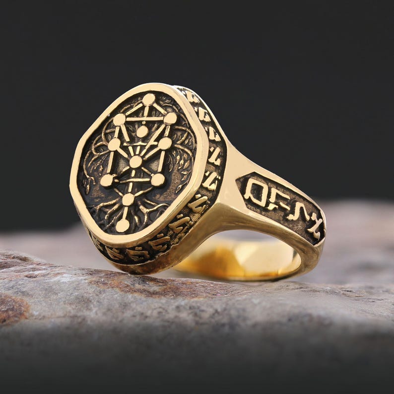 Op de afbeelding: Een goudkleurige ring met een ingewikkeld ontwerp. De ring heeft een gedetailleerd, verhoogd patroon op de voorkant, met een geometrisch symbool in het midden. De band heeft ge&euml;tste symbolen. De ring ligt op een stenen oppervlak.
