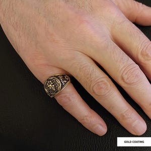 Op de afbeelding: Een vergulde ring met een zwart en gouden ontwerp. De ring heeft een cirkelvormig ontwerp met ingewikkelde details. De ring wordt gedragen aan een vinger.