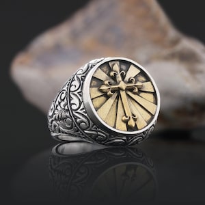 May include: A silver ring with a gold cross design. The cross is surrounded by a sunburst pattern. The band of the ring is decorated with an intricate floral design.