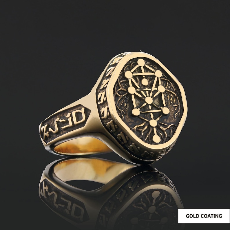 Op de afbeelding: Een goudkleurige ring met een gegraveerd ontwerp met een geometrisch patroon en symbolen. De ring heeft een rechthoekige voorkant met afgeronde randen en gedetailleerde zijgravures. De woorden "GOLD COATING" zijn zichtbaar.