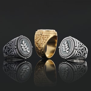 Könnte beinhalten: Drei Herrenringe, zwei silber und ein gold. Die silbernen Ringe haben ein Blattdesign und der goldene Ring hat ein filigraneres Design.