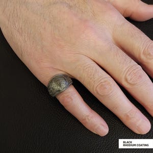 Op de afbeelding: Een zilveren ring met een gedetailleerd ontwerp aan de bovenkant. De ring zit aan een vinger en heeft een zwarte rhodium coating.