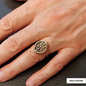 Op de afbeelding: Goudkleurige ring met een vierkante voorkant met een geometrisch ontwerp. De ring heeft een sierlijke rand en wordt om een vinger gedragen. De tekst "GOLD COATING" is rechtsonder zichtbaar.