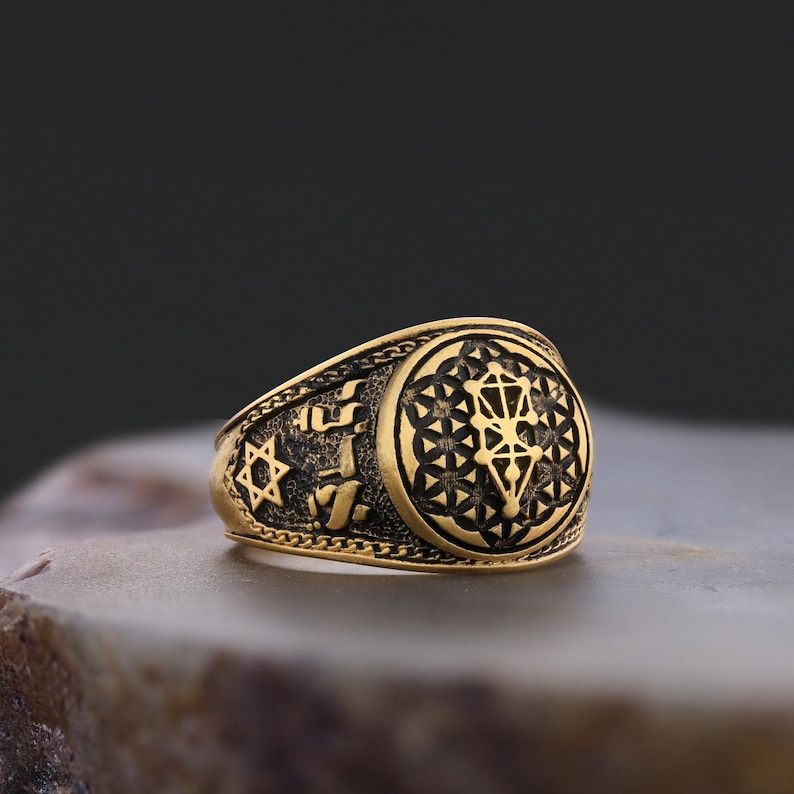 Op de afbeelding: Een gouden ring met een zwart en gouden ontwerp. De ring heeft een Davidster, Hebreeuwse tekst en een geometrisch patroon.