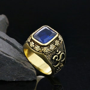 Może przedstawiać: Złoty pierścionek z niebieskim kamieniem szlachetnym i skomplikowanym wzorem. Pierścionek zawiera symbol mantry Om.