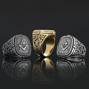 Könnte beinhalten: Drei silberne Freimaurer-Ringe mit gravierten Symbolen. Der Ring links und rechts hat ein Quadrat- und Zirkel-Design mit einer Krone. Der Ring in der Mitte ist golden mit einem Fleur-de-lis-Design.