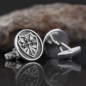 Puede incluir: Gemelos de plata con un diseño redondo que presenta un escudo con una cruz y acentos de flor de lis.