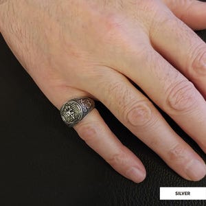 Op de afbeelding: Een zilveren ring met een gedetailleerd ontwerp met een cirkelvormig middelpunt. De ring wordt aan een vinger gedragen.