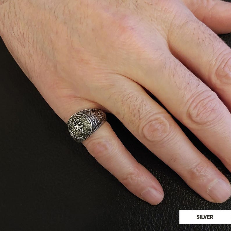 Op de afbeelding: Een zilveren ring met een gedetailleerd ontwerp met een cirkelvormig middelpunt. De ring wordt aan een vinger gedragen.