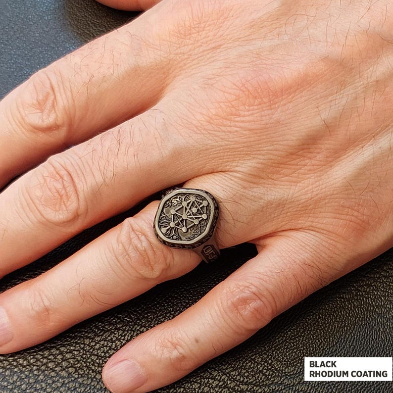 Op de afbeelding: Een zwart rodium-gecoate ring met een gedetailleerd geometrisch ontwerp. De ring heeft een vierkante voorkant met een ingewikkeld patroon en wordt aan een hand gedragen. De tekst "Black Rhodium Coating" is zichtbaar.