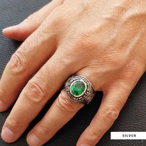 Könnte beinhalten: Ein silberner Ring mit einem grünen Edelstein und einer Inschrift um das Band. Der Ring wird an einem Finger getragen.