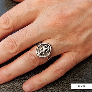 Op de afbeelding: Zilveren ring met een gedetailleerd, geometrisch ontwerp op een vierkante voorkant. De ring heeft een donkere, getextureerde achtergrond met een lichter zilveren ontwerp in het midden. De band is koperkleurig.