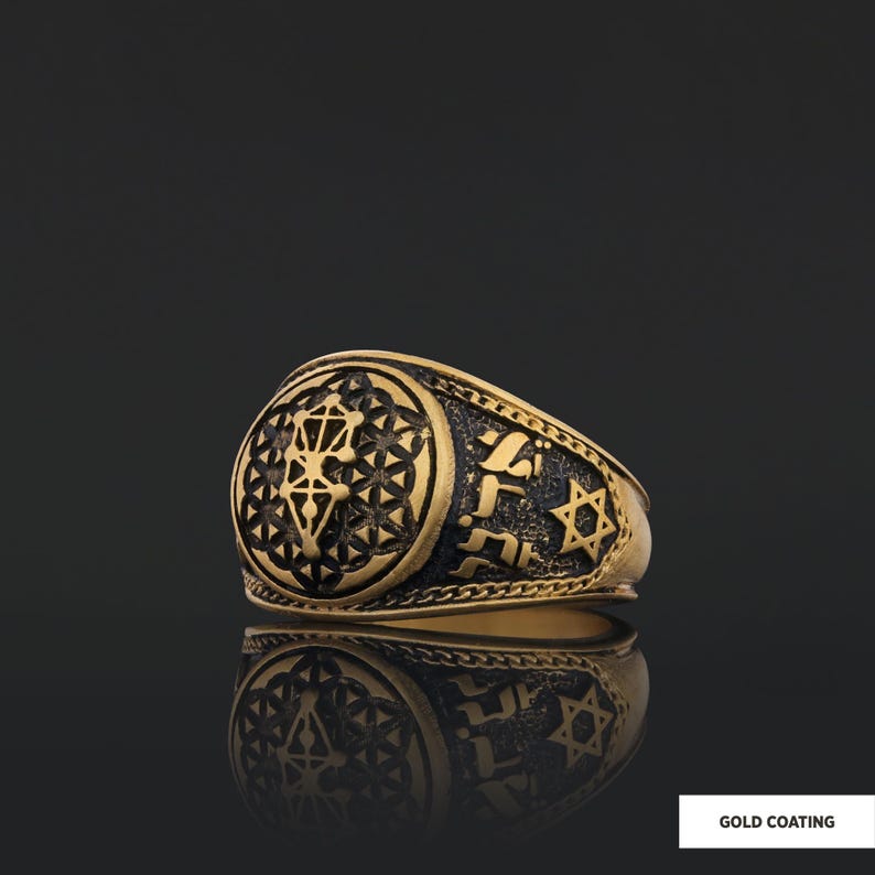Op de afbeelding: Een gouden ring met een zwart emaille ontwerp met een geometrisch patroon, Hebreeuwse tekst en een Davidster. De ring heeft een brede band en een verhoogd ontwerp.