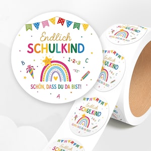 Könnte beinhalten: Eine Rolle runder weißer Aufkleber mit einem bunten Design aus Regenbogen, lächelndem Stern, Wimpelkette, Stift und dem Text Endlich SCHULKIND SCHÖN, DASS DU DA BIST, ergänzt durch Buchstaben und Zahlen auf weißem Hintergrund.